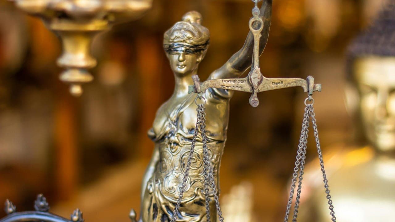 Detailaufnahme einer metallenen Justitia-Figur mit verbundenen Augen und Waage, vor unscharfem goldenem Hintergrund.