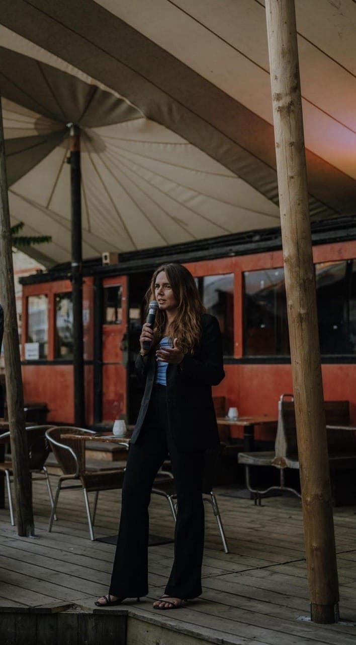 Rechtsanwältin Diederichs spricht bei einer Veranstaltung im Freien. Sie steht auf einer Holzterrasse unter großen Sonnensegeln und hält ein Mikrofon, während sie vor einem roten Waggon im Hintergrund referiert.